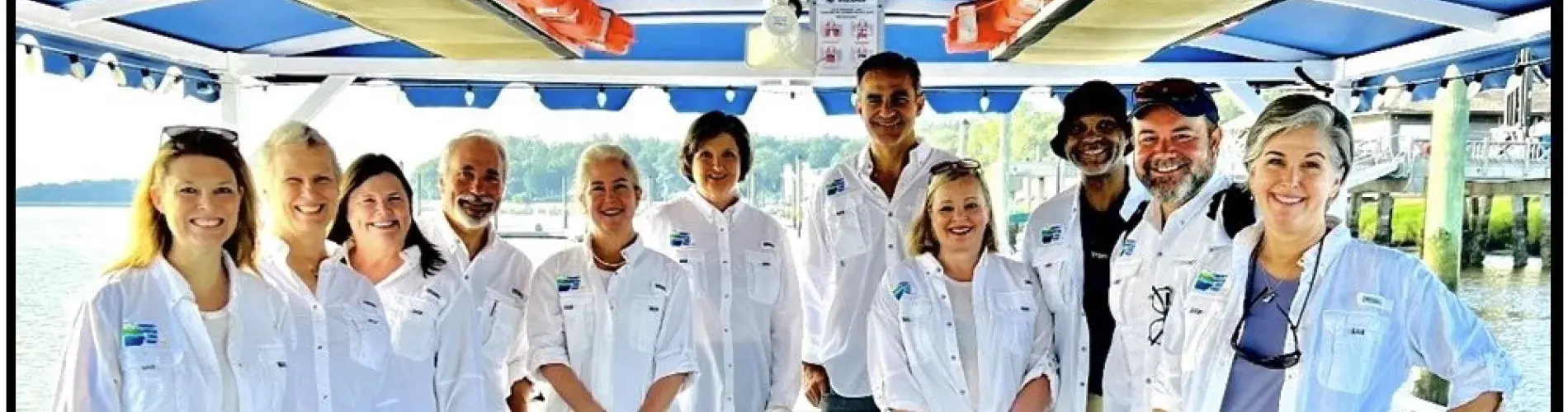 Group of people on a boat, wearing white shirts, smiling under a blue-striped canopy.