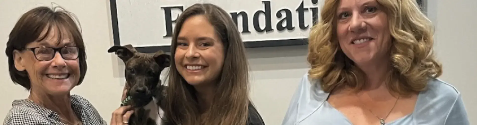 Women smiling with a small dog, standing in front of a foundation sign.